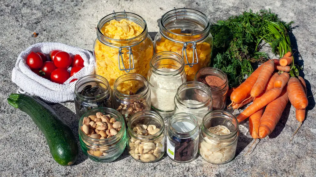 Frascos de vidrio con pasta, frutos secos, harina y especias a granel junto a zanahorias, tomates y calabacín